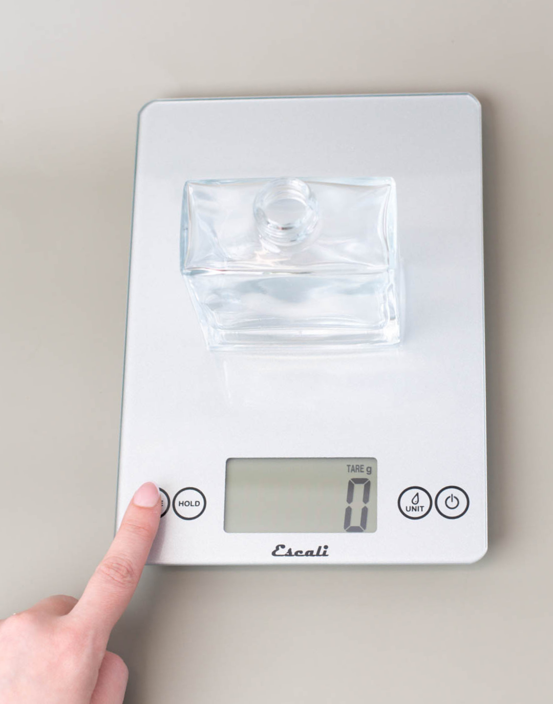 A hand pressing tare on a scale with an empty glass reed diffuser on it to set the scale to 0.